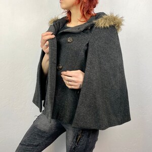 May include: A charcoal gray cape coat with a hood trimmed with faux fur. The coat features a double-breasted button closure and a classic cape design. The coat is made of a soft, wool-like material.