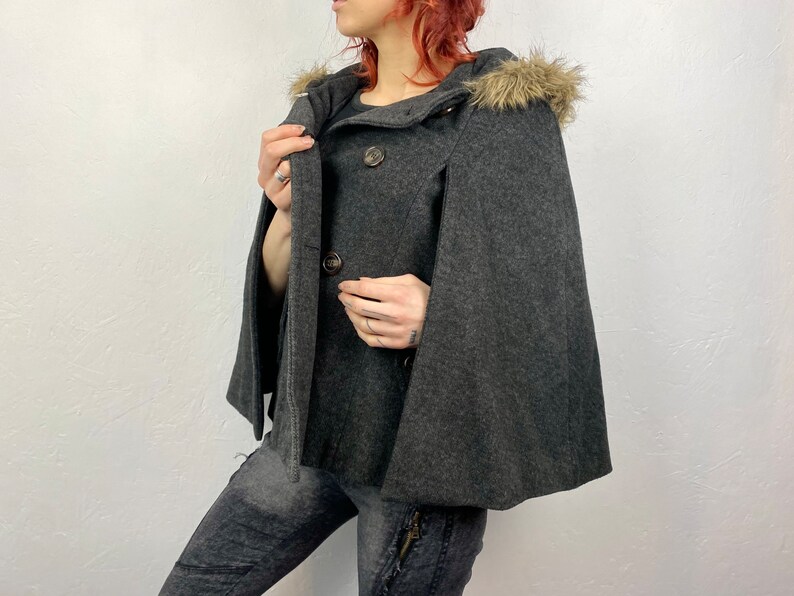 May include: A charcoal gray cape coat with a hood trimmed with faux fur. The coat features a double-breasted button closure and a classic cape design. The coat is made of a soft, wool-like material.