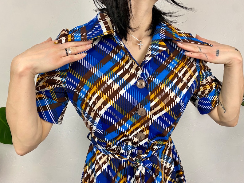 May include: A blue, brown, and white plaid dress with a collared neckline, short sleeves, and a button-up front. The dress has a belt at the waist.
