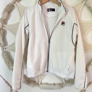 May include: White Fila track jacket with black trim and a full zipper. The jacket features the Fila logo on the chest and a small logo on the collar. The jacket is hanging on a wooden hanger.