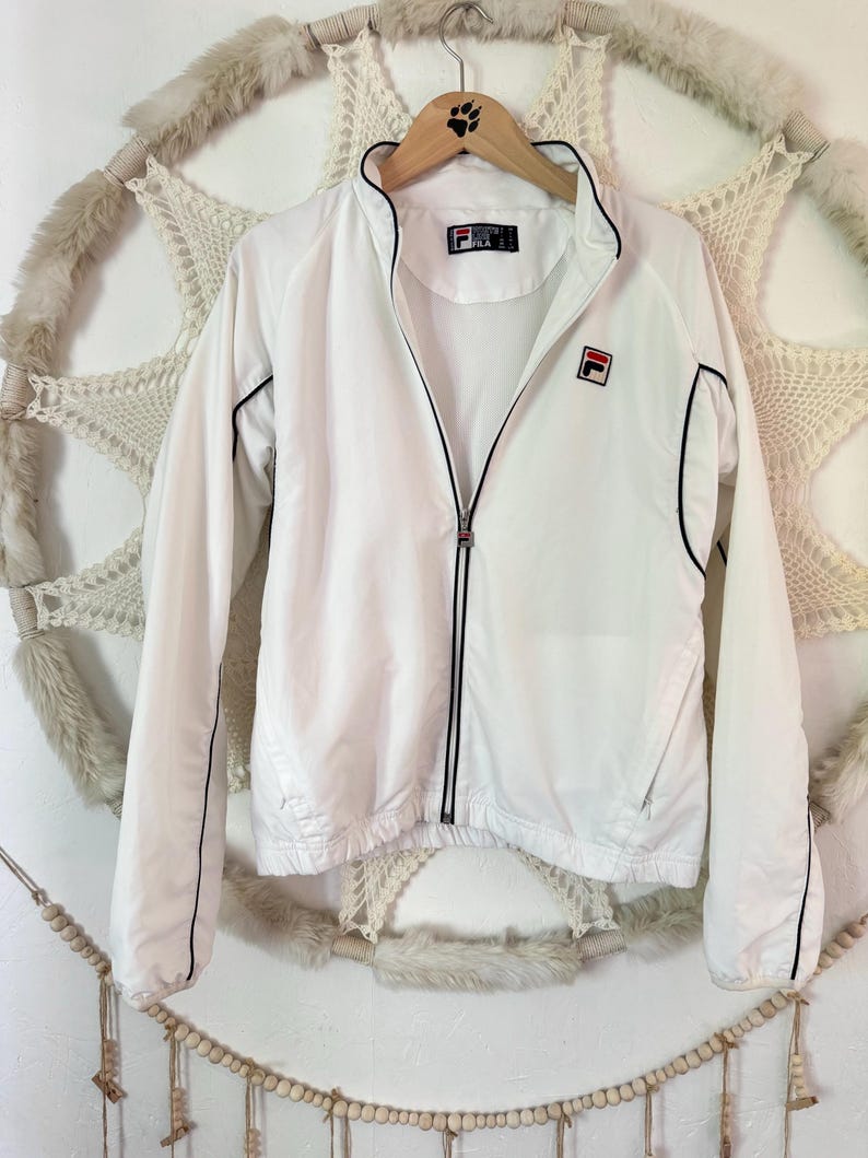 May include: White Fila track jacket with black trim and a full zipper. The jacket features the Fila logo on the chest and a small logo on the collar. The jacket is hanging on a wooden hanger.