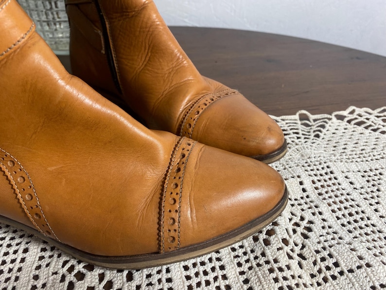 K&ouml;nnte beinhalten: Ein Paar hellbraune Leder-Ankle Boots. Die Stiefel haben eine spitze Zehenpartie mit dekorativen Perforationen und einen seitlichen Rei&szlig;verschluss. Das Leder hat ein abgenutztes Aussehen. Die Stiefel stehen auf einer Holzoberfl&auml;che.