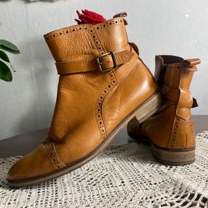 K&ouml;nnte beinhalten: Ein Paar hellbraune Leder-Ankle Boots mit Schnallenriemen und dekorativen Perforationen. Die Stiefel haben einen niedrigen Absatz und eine runde Spitze. Das Leder hat ein abgenutztes Aussehen. Die Stiefel stehen auf einem Tisch.