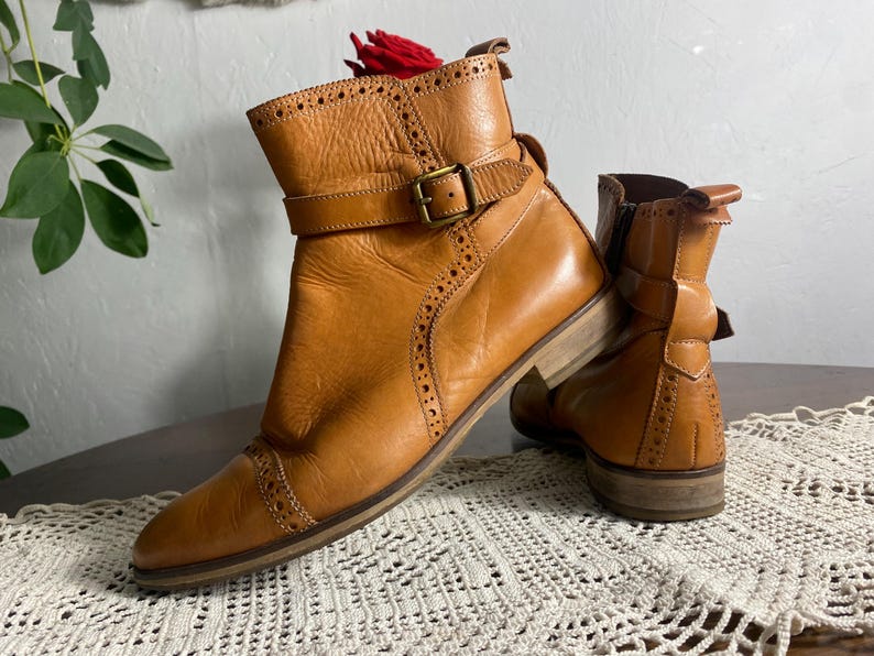 K&ouml;nnte beinhalten: Ein Paar hellbraune Leder-Ankle Boots mit Schnallenriemen und dekorativen Perforationen. Die Stiefel haben einen niedrigen Absatz und eine runde Spitze. Das Leder hat ein abgenutztes Aussehen. Die Stiefel stehen auf einem Tisch.