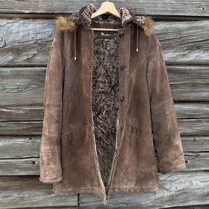 May include: Brown suede coat with a fur-lined hood and interior. The coat has a button-up front, side pockets, and a label that reads "Mellisa". The coat is hanging on a wooden hanger against a wooden background.