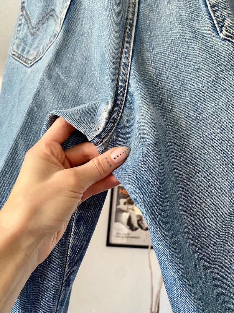 May include: Light blue denim jeans with a classic five-pocket design. The jeans show signs of wear and tear, with visible fading and distressing. The image highlights the texture and details of the denim fabric, showcasing the stitching and pocket design.
