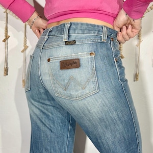 May include: Light blue denim Wrangler jeans. The jeans have a classic five-pocket design, a brown leather patch with the Wrangler logo, and a faded wash. The back pocket features the iconic Wrangler "W" stitching.