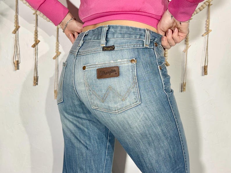 May include: Light blue denim Wrangler jeans. The jeans have a classic five-pocket design, a brown leather patch with the Wrangler logo, and a faded wash. The back pocket features the iconic Wrangler "W" stitching.