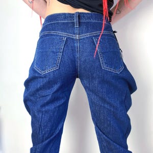 Può includere: Un paio di jeans in denim blu scuro con una vestibilità rilassata. I jeans hanno una gamba dritta e una lunghezza leggermente corta.