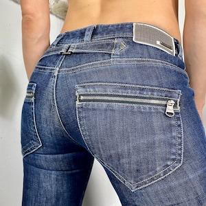 May include: Dark blue denim jeans with a zippered pocket on the back. The jeans have a belt loop and a silver buckle.