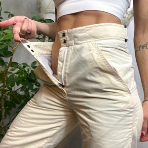May include: Cream-coloured ski trousers with a high waist and a zip closure. The trousers have a quilted texture and feature pockets on the sides. The image shows the trousers unbuttoned and unzipped, revealing the interior lining.