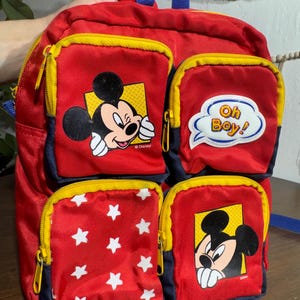 May include: Red and blue Mickey Mouse backpack with four zippered pockets. The pockets feature Mickey Mouse graphics and white stars on a red background. The backpack has yellow zippers and a blue handle.