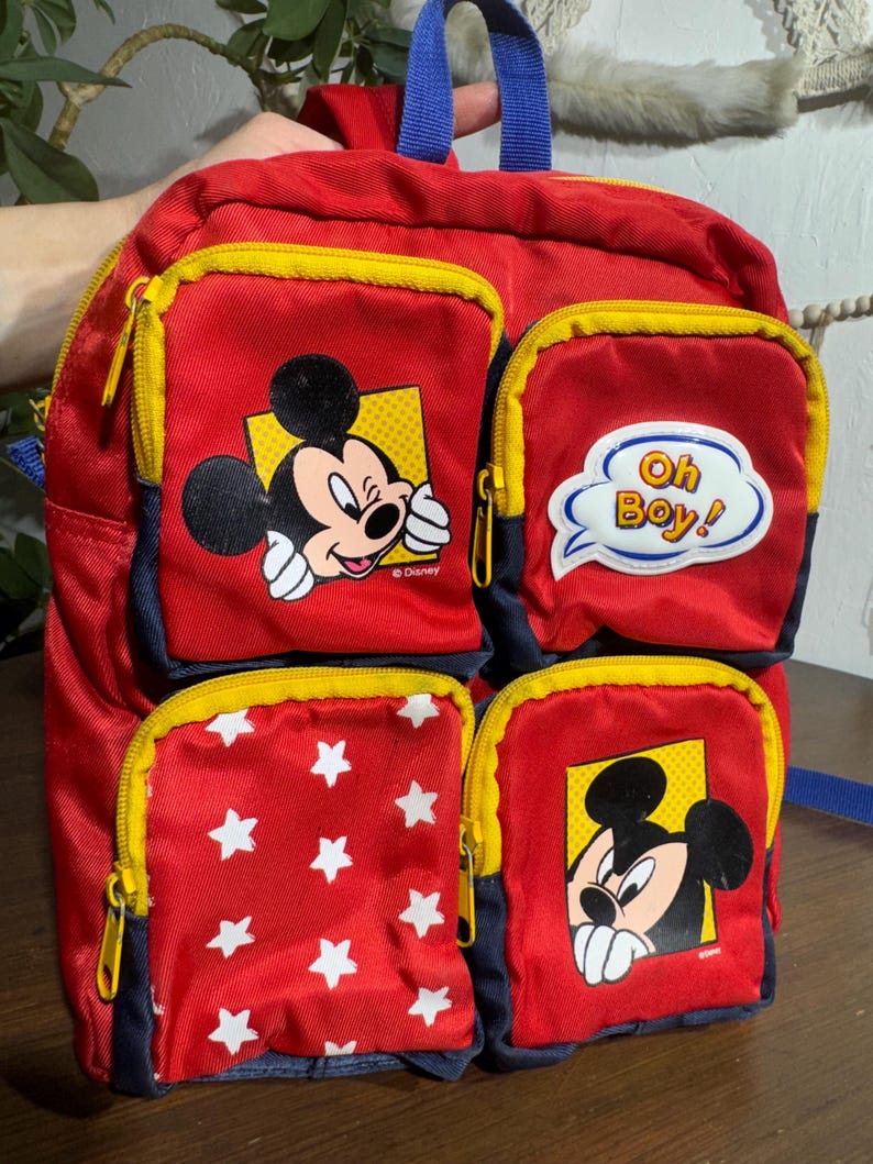 May include: Red and blue Mickey Mouse backpack with four zippered pockets. The pockets feature Mickey Mouse graphics and white stars on a red background. The backpack has yellow zippers and a blue handle.