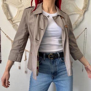 May include: A beige jacket with three-quarter sleeves and a double-breasted design. The jacket has a collared neckline and is worn over a white top and blue jeans. A black belt with a gold buckle is visible.
