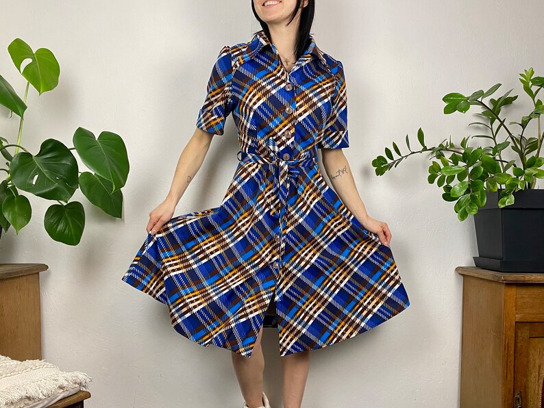 May include: A blue, white, and brown plaid dress with a button-up front and a belt. The dress has a short sleeve and a flared skirt.