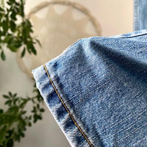 May include: Close-up of light blue denim fabric with visible texture and a contrasting yellow-brown stitch along the edge. The denim appears to be from a pair of jeans, showcasing the material's weave and color.