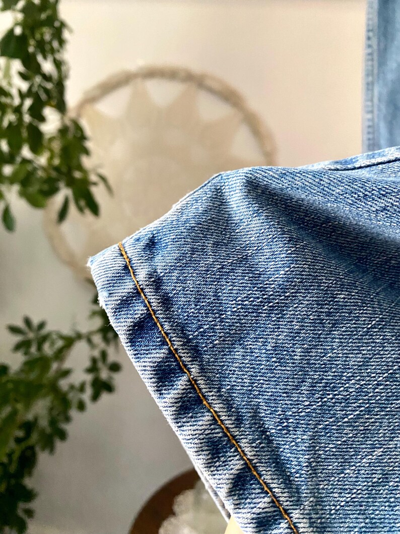 May include: Close-up of light blue denim fabric with visible texture and a contrasting yellow-brown stitch along the edge. The denim appears to be from a pair of jeans, showcasing the material's weave and color.