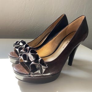 Könnte beinhalten: Braune Peeptoe-Heels mit einem gerafften Stoffdetail an der Vorderseite. Die Absätze sind etwa 10 cm hoch und haben einen dünnen Riemen um den Knöchel. Die Schuhe sind mit "Melani" gekennzeichnet.