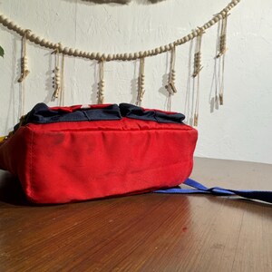 May include: A red backpack with a blue strap and accents. The bag has two small pockets on top and a white star design. The bag is sitting on a wooden surface.