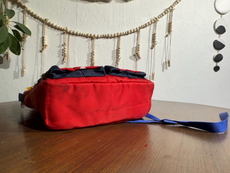May include: A red backpack with a blue strap and accents. The bag has two small pockets on top and a white star design. The bag is sitting on a wooden surface.