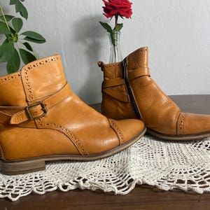 K&ouml;nnte beinhalten: Ein Paar hellbraune Leder-Ankle-Boots mit Ziern&auml;hten und Schnallen. Ein Stiefel hat einen seitlichen Rei&szlig;verschluss. Die Stiefel werden auf einer wei&szlig;en Spitzendecke mit einer roten Rose in einer Vase im Hintergrund pr&auml;sentiert.