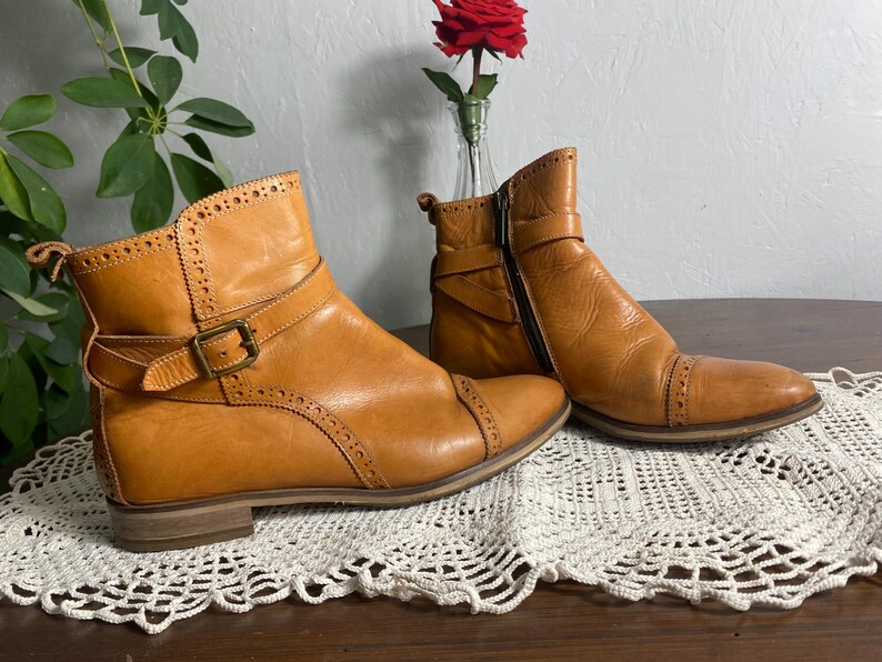 K&ouml;nnte beinhalten: Ein Paar hellbraune Leder-Ankle-Boots mit Ziern&auml;hten und Schnallen. Ein Stiefel hat einen seitlichen Rei&szlig;verschluss. Die Stiefel werden auf einer wei&szlig;en Spitzendecke mit einer roten Rose in einer Vase im Hintergrund pr&auml;sentiert.