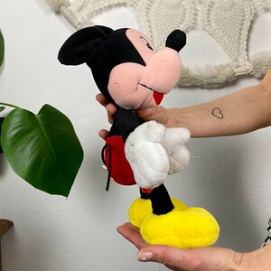 May include: A plush Mickey Mouse toy with black ears, a red and white shirt, and yellow shoes. The toy is being held by a person's hand.