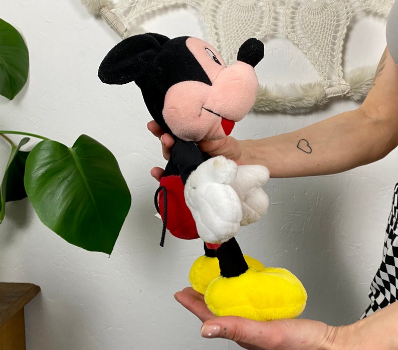 May include: A plush Mickey Mouse toy with black ears, a red and white shirt, and yellow shoes. The toy is being held by a person's hand.
