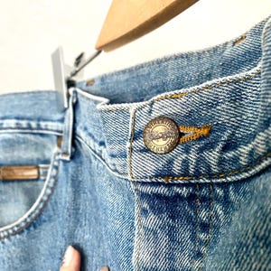 May include: Close-up of light blue denim jeans with a metal button that reads "Authentic". The jeans are hanging on a wooden hanger. The jeans have a classic five-pocket design.