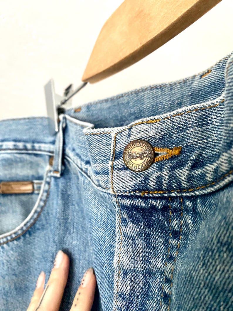 May include: Close-up of light blue denim jeans with a metal button that reads "Authentic". The jeans are hanging on a wooden hanger. The jeans have a classic five-pocket design.