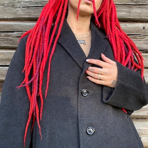 Può includere: Un cappotto di lana grigio scuro con tre bottoni neri. Il cappotto è indossato sopra un maglione beige. La persona ha dreadlocks rosso vivo con riflessi viola. Lo sfondo è un muro di legno.