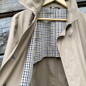 Può includere: Un trench coat beige dal design classico, con ampio colletto e bottoni. La fodera interna presenta un motivo a quadri neri, bianchi e verde oliva. Il cappotto è appeso a un'appendiabiti di legno.