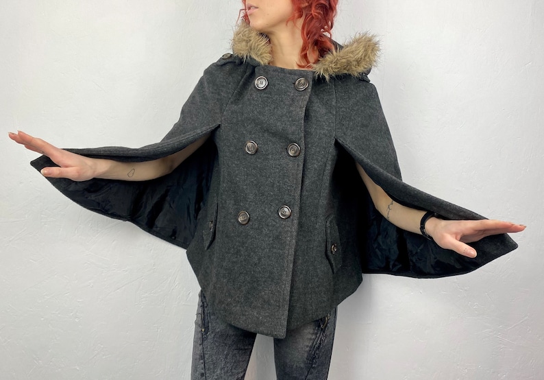 May include: A gray cape coat with a faux fur-lined hood and double-breasted button closure. The coat has a cape design with arm openings and two front pockets. The lining is black.