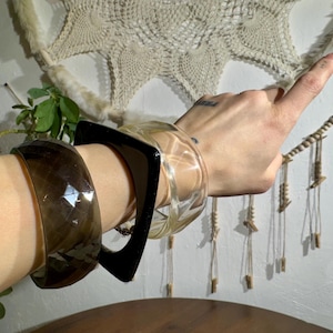 Puede incluir: Una colección de pulseras de varios estilos. Una es una pulsera gruesa facetada de color marrón oscuro. Otra es una pulsera transparente de forma cuadrada. La tercera es una pulsera transparente y redondeada. Las pulseras se llevan en la muñeca.