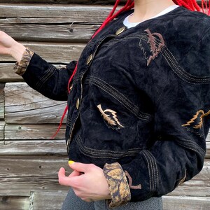 May include: A black suede jacket with gold stitching and brown leaf embroidery. The jacket has a button closure and a cropped style.