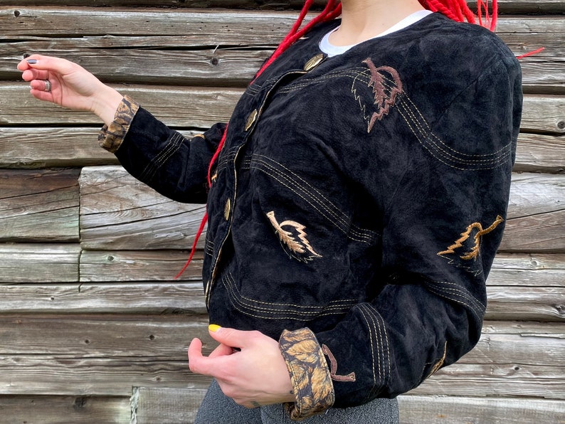 May include: A black suede jacket with gold stitching and brown leaf embroidery. The jacket has a button closure and a cropped style.