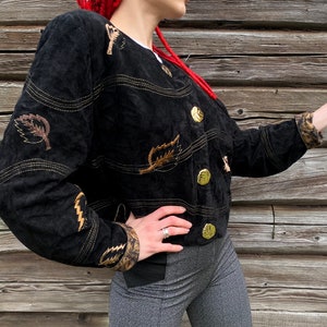 May include: A black suede jacket with gold buttons and leaf embroidery. The jacket has a cropped fit and a relaxed silhouette.