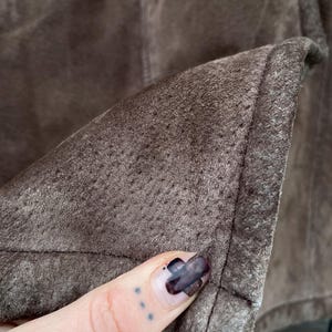 May include: Close-up of a section of brown suede fabric. The material has a textured, porous surface and is stitched along the edges. The color is a muted, earthy brown.