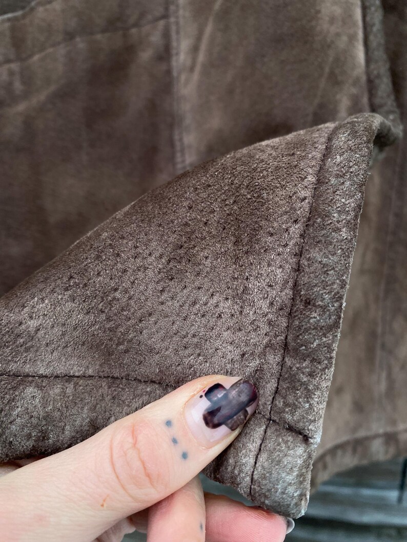 May include: Close-up of a section of brown suede fabric. The material has a textured, porous surface and is stitched along the edges. The color is a muted, earthy brown.