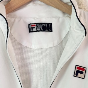 May include: White FILA jacket with a black trim and a full zipper. The jacket features the FILA logo on the chest and a label that reads "WHITE LINE FILA". The jacket is hanging on a wooden hanger.
