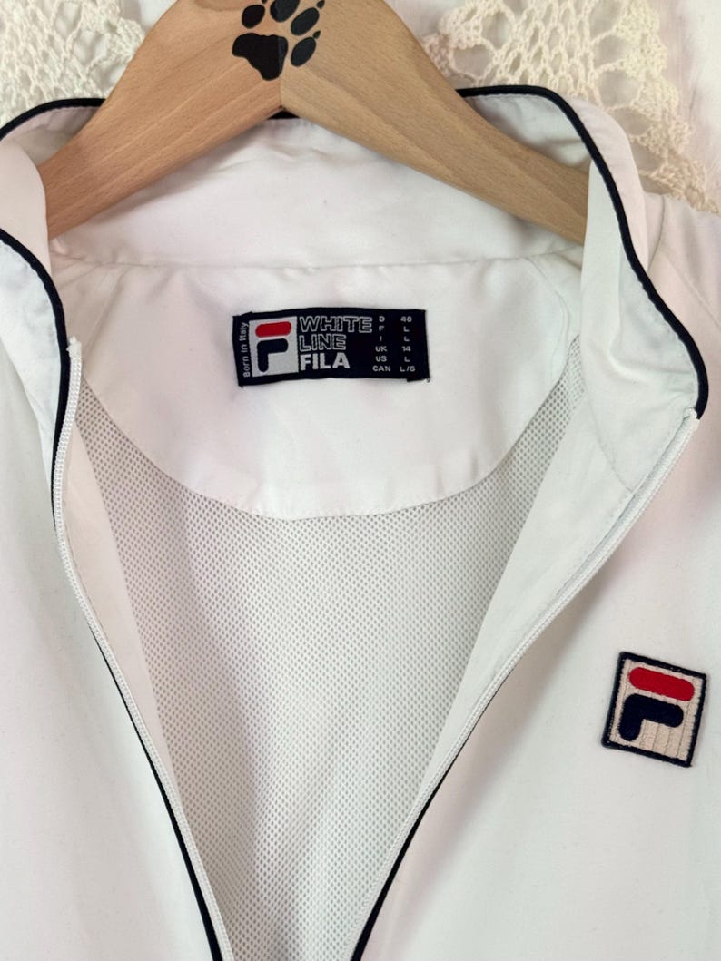 May include: White FILA jacket with a black trim and a full zipper. The jacket features the FILA logo on the chest and a label that reads "WHITE LINE FILA". The jacket is hanging on a wooden hanger.