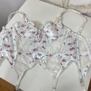 Könnte beinhalten: Weiße florale Spitzen-Dessous mit Satin-Cups und verstellbaren Trägern. Die Dessous haben ein Korsett-ähnliches Oberteil und Strumpfhalter-Bänder.