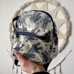 Peut inclure: Un petit sac à dos crème avec un motif répété de robots gris. Le sac à dos a une fermeture éclair et des bretelles bleu marine. Le sac est tenu par une main.
