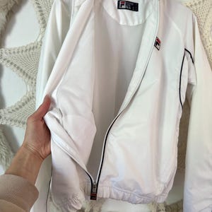 May include: White FILA track jacket with black trim and a full zipper. The jacket features the FILA logo on the chest and a tag on the collar. The jacket is hanging on a wooden hanger.
