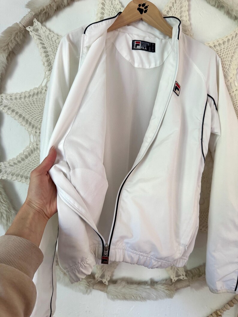 May include: White FILA track jacket with black trim and a full zipper. The jacket features the FILA logo on the chest and a tag on the collar. The jacket is hanging on a wooden hanger.