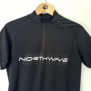 Pode incluir: Uma camisola de ciclismo preta de manga curta com gola alta e fecho frontal. O peito apresenta o texto branco NORTHWAVE com um efeito de sombra repetitivo. Um pequeno logótipo vermelho, amarelo e preto é visível na bainha da manga esquerda.