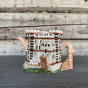 May include: A white ceramic teapot shaped like a castle with brown accents and a brown handle. The teapot has a detailed design with windows and a gate.