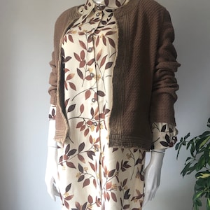 Op de afbeelding: Een bruine wafel gebreide cardigan met een rafelige zoom, gedragen over een wit overhemd met knopen en een bruine bladprint.