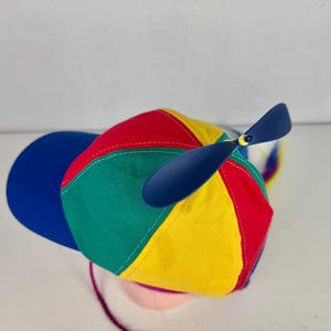 Può includere: Un cappellino da baseball multicolore con pannelli in tessuto rosso, giallo, verde e blu. Il cappellino presenta una visiera blu tinta unita e un'elica in plastica blu scuro fissata sulla parte superiore, con una nappa arcobaleno che pende dal retro.
