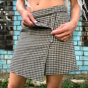 Puede incluir: Una minifalda de pata de gallo en blanco y negro con un detalle de cremallera. La falda es plisada y tiene una silueta ajustada.